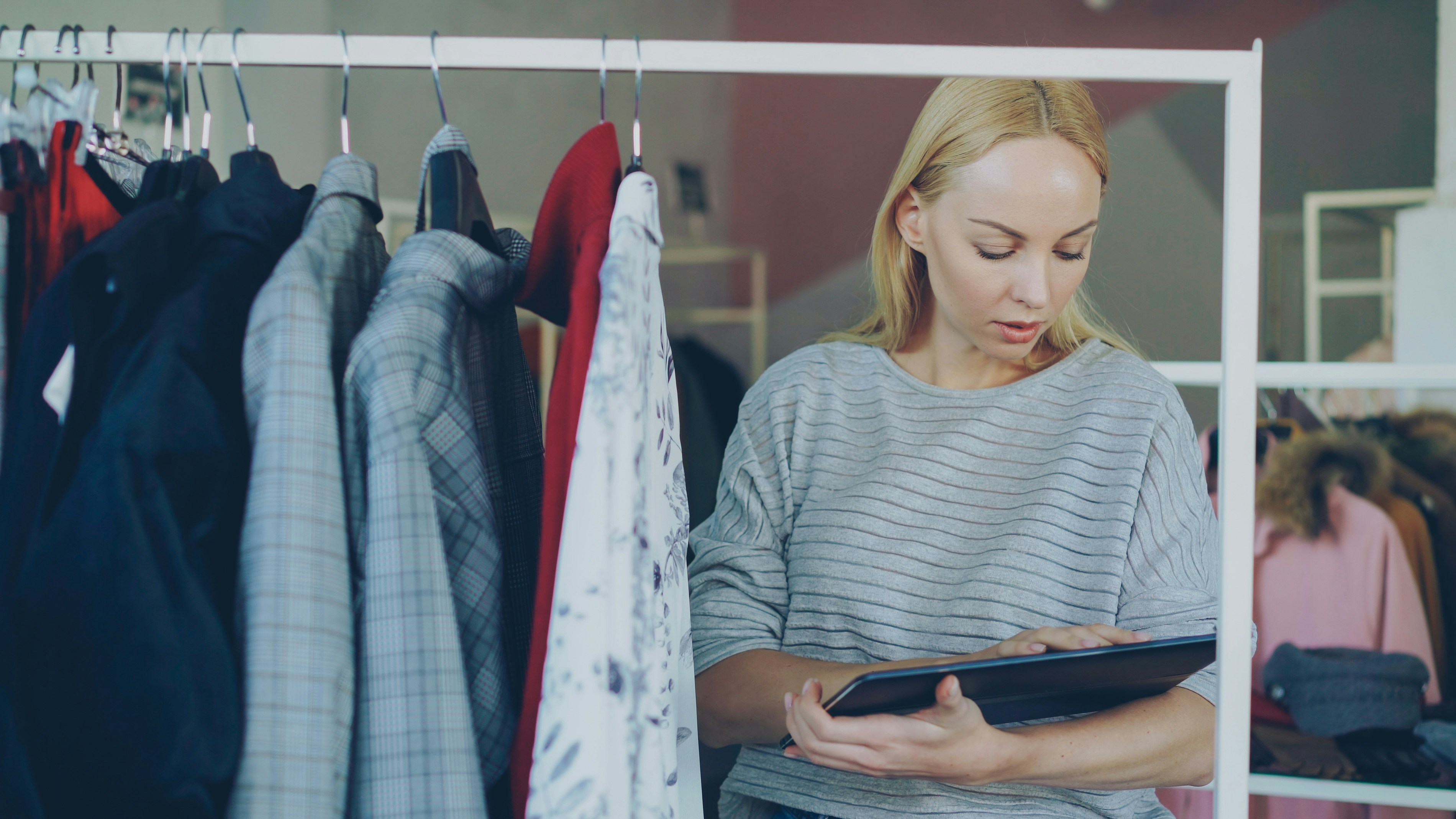 A woman in a striped sweater holding a tablet in a small clothing boutique, focused and reviewing stock.
